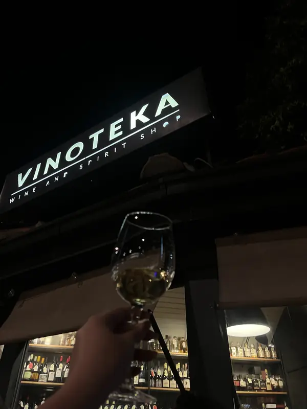 Čaša belog vina podignuta ispod osvetljene table VINOTEKA